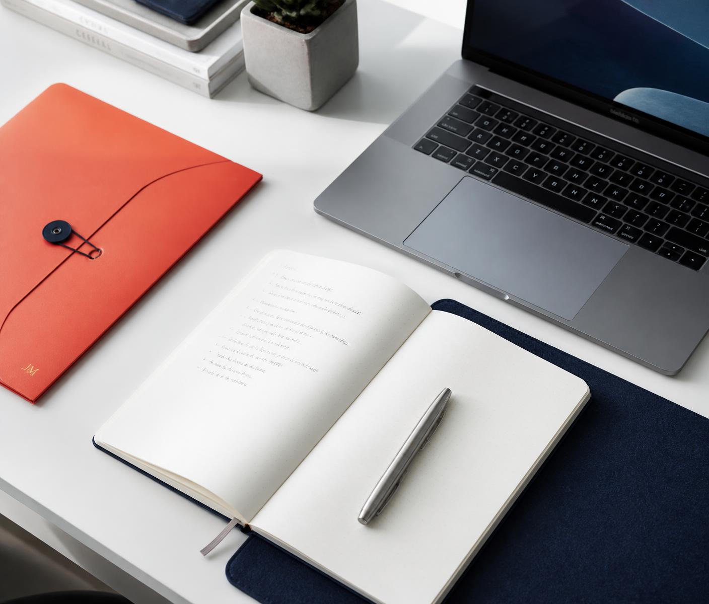 White desk with an open notebook, laptop, navy desk mat, and a coral folder